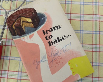 May include: A vintage cookbook titled "Learn to Bake..." with a retro illustration of a chocolate cake on a plate. The cover is pink and white with the words "You'll Love it!" written in cursive. A red-handled whisk is in the background.