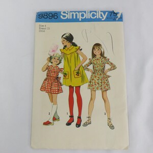 Puede incluir: Patrón de costura Simplicity vintage 9896 para vestidos de niña. La portada del patrón presenta ilustraciones de tres niñas con diferentes vestidos. Los vestidos incluyen un vestido amarillo con bolsillos de cerezas, un vestido a cuadros rojos y blancos y un vestido con estampado floral.