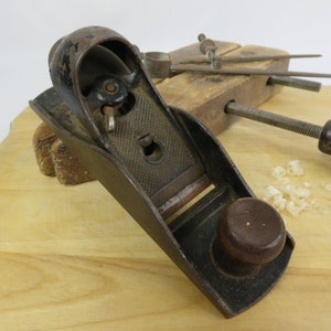 May include: A vintage hand plane with a dark metal body and a wooden knob. The plane rests on a light wood surface, with wood shavings nearby. Other woodworking tools are in the background.