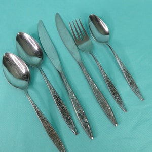 May include: A set of six stainless steel flatware pieces, including spoons, knives, and a fork. The handles have a textured geometric pattern. The flatware is arranged on a light blue surface, showcasing a vintage or retro style.