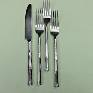 Könnte beinhalten: Ein Edelstahl-Besteckset mit einem Messer und drei Gabeln. Die Griffe haben ein bambusähnliches Design. Die Zinken der Gabeln sind sichtbar. Das Besteck befindet sich vor einem hellgrünen Hintergrund.