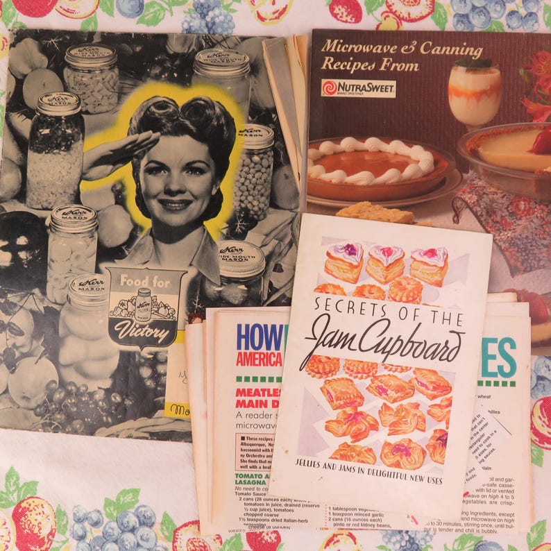 May include: A collection of vintage cookbooks and recipe pamphlets. The image features a black and white illustration of a woman saluting, surrounded by jars of preserved food. Titles include "Food for Victory" and "Secrets of the Jam Cupboard."