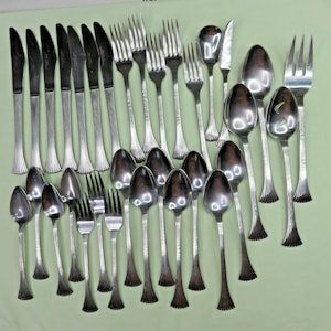 Op de afbeelding: Een verzameling vintage bestek, waaronder messen, vorken en lepels, gerangschikt op een lichtgroen oppervlak. Het bestek heeft een glanzende, metalen afwerking met decoratieve handvatten. De set bevat verschillende maten lepels en vorken.