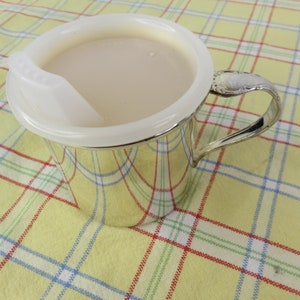 Könnte beinhalten: Ein silberfarbener Babybecher mit weißem Kunststoffdeckel und Griff. Der Becher ist mit einer hellfarbigen Flüssigkeit gefüllt. Der Griff hat ein dekoratives Design. Der Becher steht auf einer gelben karierten Tischdecke.