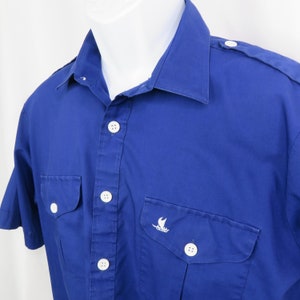 Puede incluir: Camisa azul cobalto de manga corta con dos bolsillos en el pecho y botones blancos. La camisa presenta un pequeño emblema de velero blanco en un bolsillo y charreteras en los hombros. El cuello es de estilo clásico.