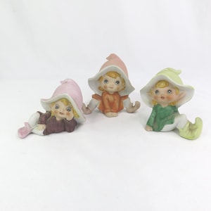 May include: Three ceramic elf figurines with blonde hair and blue eyes. Each elf wears a different coloured outfit and hat: pink, orange, and green. The elves are in various poses, with one lying down and the others sitting.
