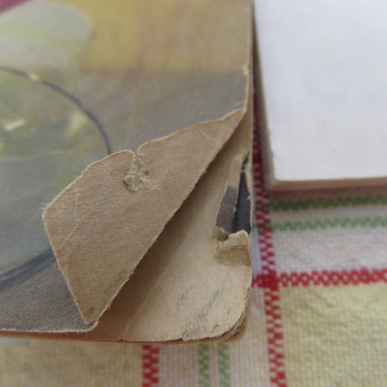 May include: Close-up of a vintage book's damaged corner. The cover is peeling away, revealing a tan, textured surface with a faded image. The edges are worn, showing signs of age and use. The book's pages are visible.