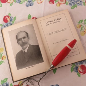 May include: An open vintage cookbook titled "Good Food How to Prepare It" with a black and white portrait of George E. Cornforth. A red and silver whisk rests on the book, with a floral tablecloth in the background.
