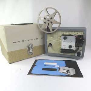 May include: A vintage Kodak Brownie 8 movie projector in gray with a metal film reel, a beige and tan carrying case labeled Brownie 8, and a black and blue instruction manual featuring the text Brownie 8 movie projector model 10.