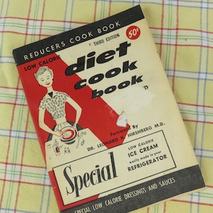 May include: Vintage "Diet Cook Book" with a red and black cover, featuring a retro illustration of a woman cooking. The book is labeled "Low Calorie" and includes text about low-calorie ice cream and dressings.