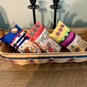 May include: A rectangular basket with leather handles holds four decorative, handmade fabric sachets. The sachets are in various colors and patterns, including blue, floral, and yellow, with embroidered details and button embellishments. The basket sits on a wooden surface.