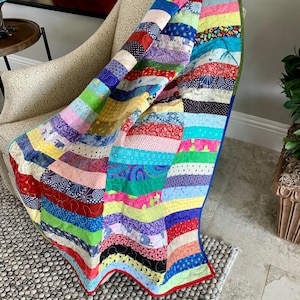 May include: A colorful patchwork quilt with stripes of various colors and patterns, including red, blue, green, yellow, and purple. The quilt is folded and draped over a chair with a light brown fabric.