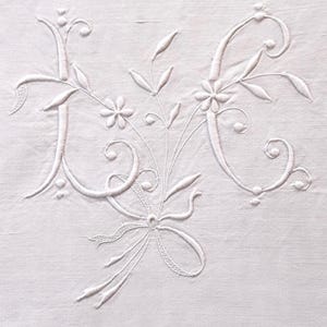 May include: A white textile featuring intricate white embroidery of the letters L and C intertwined with floral motifs, delicate stems, and a central bow. The embroidery uses raised satin stitching and fine line work on a plain white fabric background.