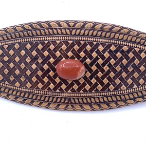 May include: Oval-shaped wooden hair barrette with a woven pattern and a decorative border. The barrette features a reddish-brown stone in the center. The wood has a dark brown and tan color scheme.