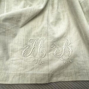 May include: Close-up of a light beige linen fabric with embroidered initials "A B" in a decorative script. The embroidery is in a matching off-white color, with floral accents.