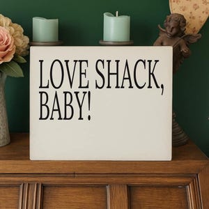 Könnte beinhalten: Ein cremefarbenes rechteckiges Schild mit dem Text "LOVE SHACK, BABY!" in Schwarz. Das Schild steht auf einer Holzoberfläche, mit zwei türkisfarbenen Kerzen und einer Vase mit rosa Rosen im Hintergrund.
