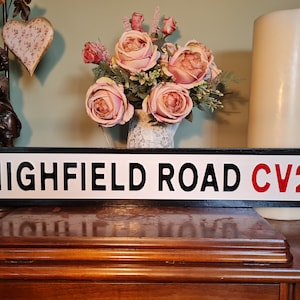 Può includere: Un cartello stradale bianco e nero con il testo "HIGHFIELD ROAD CV2" in lettere bianche su sfondo nero. Il cartello ha bordi arrotondati e un bordo nero. Il cartello è su una superficie di legno.