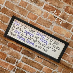 May include: A framed sign with the text "Blue is the colour, football is the game..." in blue and black. The sign is set against a brick wall background.