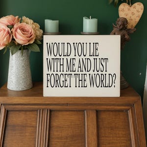 Pode incluir: Um armário de madeira exibe uma placa creme com texto preto: "WOULD YOU LIE WITH ME AND JUST FORGET THE WORLD?" Duas velas verde-azuladas estão em cima da placa. Um vaso de rosas cor-de-rosa e um anjo decorativo também estão presentes.
