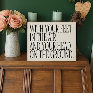May include: A cream-coloured wooden sign with the quote "WITH YOUR FEET IN THE AIR AND YOUR HEAD ON THE GROUND" in black lettering. The sign is on a wooden surface, with a vase of pink roses and two green candles in the background.