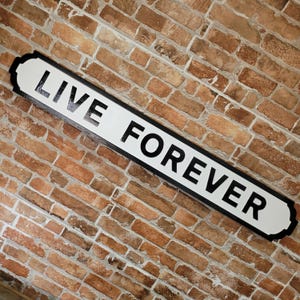 Puede incluir: Un letrero rectangular con las palabras "LIVE FOREVER" en letras mayúsculas negras y en negrita sobre un fondo blanco. El letrero tiene un borde negro y está montado en una pared de ladrillo.