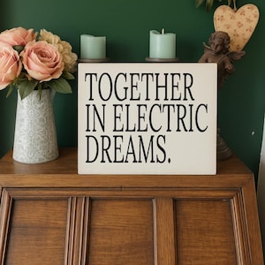 Può includere: Un mobile in legno espone un cartello color crema con la scritta "TOGETHER IN ELECTRIC DREAMS". Due candele turchesi sono sopra il cartello. Sono presenti anche un vaso di rose rosa e una statua di angelo decorativa.