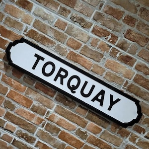 May include: A white street sign with the word "TORQUAY" in black lettering. The sign has a black border and is mounted on a brick wall with varying shades of brown and red bricks.