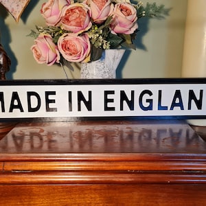 Puede incluir: Un letrero en blanco y negro con el texto "MADE IN ENGLAND" sobre un fondo blanco. El letrero está hecho de madera y tiene un borde negro.