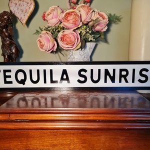 May include: A rectangular sign with the words "TEQUILA SUNRISE" in bold black letters on a white background, framed by a black border. The sign is displayed on a wooden surface, with decorative elements in the background.