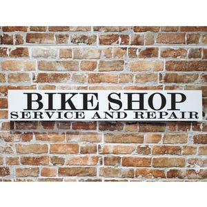 May include: A rectangular white sign with black text that reads "BIKE SHOP SERVICE AND REPAIR" is mounted on a brick wall. The bricks are various shades of brown and tan. The sign is centered and the text is in a bold, sans-serif font.
