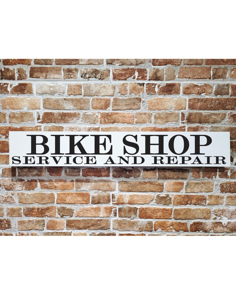 May include: A rectangular white sign with black text that reads "BIKE SHOP SERVICE AND REPAIR" is mounted on a brick wall. The bricks are various shades of brown and tan. The sign is centered and the text is in a bold, sans-serif font.
