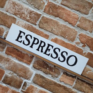 Könnte beinhalten: Ein weißes rechteckiges Schild mit dem Wort "ESPRESSO" in großen, fetten, schwarzen Buchstaben. Das Schild befindet sich vor einem Backsteinmauer-Hintergrund mit verschiedenen Braun- und Beigetönen.