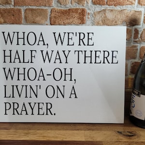 Könnte beinhalten: Ein weißes Schild mit dem Text "WHOA, WE'RE HALF WAY THERE WHOA-OH, LIVIN' ON A PRAYER." steht neben einer dunkelgrünen Champagnerflasche. Das Schild befindet sich auf einer Holzoberfläche mit einem Ziegelhintergrund.