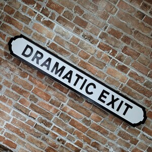 May include: A rectangular sign with a white background and a black border featuring the text DRAMATIC EXIT in bold black capital letters. The sign is mounted at an angle against a textured wall made of reddish-brown bricks with light gray mortar.