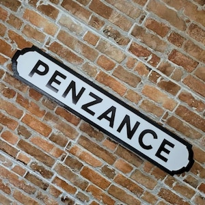 Może przedstawiać: Prostokątny znak drogowy z napisem "PENZANCE" pogrubionymi czarnymi literami na białym tle. Znak ma czarną obwódkę i jest zamontowany na ceglanej ścianie.