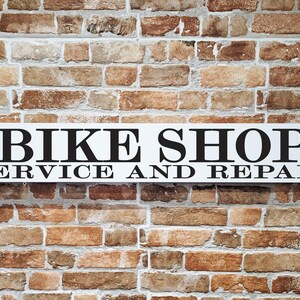 May include: A white rectangular sign with black text that reads "BIKE SHOP SERVICE AND REPAIR" is mounted on a brick wall. The brick wall has a mix of brown, tan, and reddish-brown bricks.