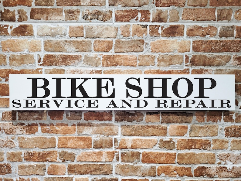 May include: A white rectangular sign with black text that reads "BIKE SHOP SERVICE AND REPAIR" is mounted on a brick wall. The brick wall has a mix of brown, tan, and reddish-brown bricks.
