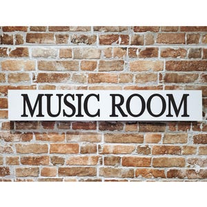 May include: A white rectangular sign displaying the words "MUSIC ROOM" in bold, black capital letters is fixed to a brick wall. The bricks are various shades of brown and tan, providing a textured backdrop.