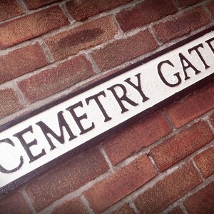 May include: A white sign with black lettering on a brick wall reads "Cemetery Gates".