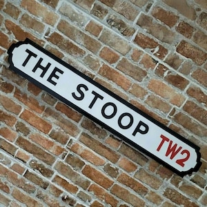 以下が含まれることがあります： 黒と赤の文字で「THE STOOP TW2」と書かれた長方形の街路標識。標識は黒い縁取りのある白い背景で、レンガの壁に取り付けられています。
