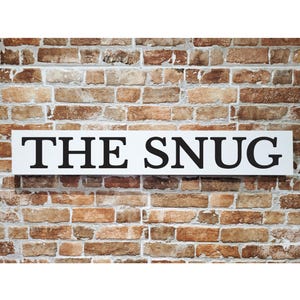Peut inclure: Une enseigne rectangulaire blanche avec les mots "THE SNUG" en lettres majuscules noires et grasses. L'enseigne est fixée sur un mur de briques aux teintes marron, beige et rouge.