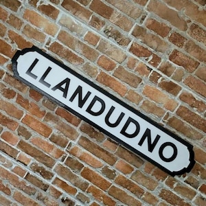 May include: A rectangular white street sign with a black border and the word LLANDUDNO printed in bold black capital letters. The sign is mounted at an angle against a wall constructed of weathered, multi-toned brown and tan bricks.