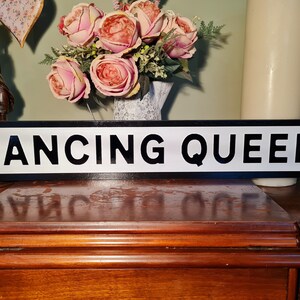 May include: Black and white sign with the text "Dancing Queen" in black letters on a white background. The sign is on a wooden surface.