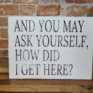 May include: A white rectangular wooden sign with black text that reads: "AND YOU MAY ASK YOURSELF, HOW DID I GET HERE?" The sign is placed on a wooden surface in front of a brick wall.