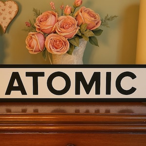May include: A black and white sign with the word "ATOMIC" in bold, black letters on a white background. The sign is framed with a black border.