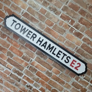 May include: A rectangular street sign with the text "TOWER HAMLETS E2" in black letters on a white background. The sign has a black border and is mounted on a brick wall.