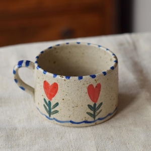 May include: A small, speckled ceramic mug with a blue handle and rim. The mug is decorated with two red heart-shaped flowers with green stems and leaves. A blue wavy line runs along the bottom of the flowers.
