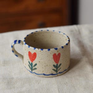 May include: A small, speckled ceramic mug with a blue handle and rim. The mug is decorated with two red heart-shaped flowers with green stems and leaves. A blue wavy line is painted at the base of the flowers.