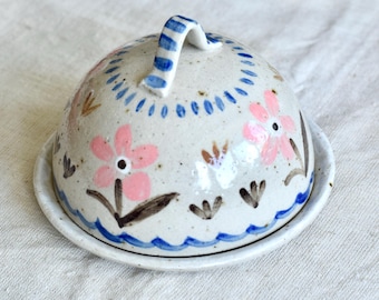 May include: A ceramic butter dish with a domed lid and a matching base. The dish is decorated with pink flowers, blue accents, and a blue and white striped handle. The design is hand-painted.