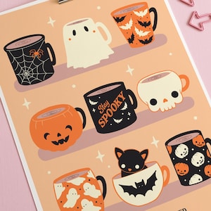 Op de afbeelding: Een Halloween-thema print met illustraties van mokken met spookachtige ontwerpen, waaronder spinnenwebben, geesten, vleermuizen en pompoenen. De zin "Stay Spooky" is zichtbaar. De print wordt vastgehouden door een zilveren clip en heeft roze pijlvormige paperclips.